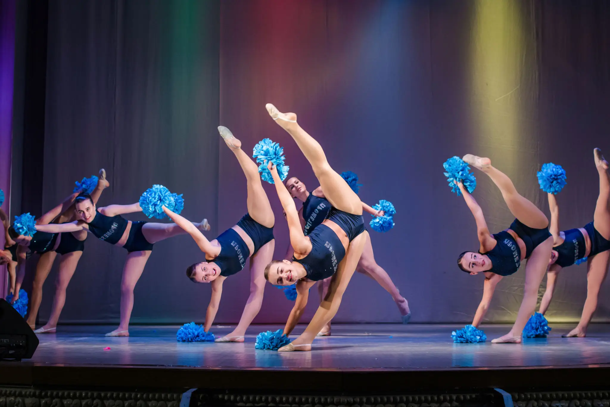 Young dancers performing a synchronized routine with blue pom-poms on stage.